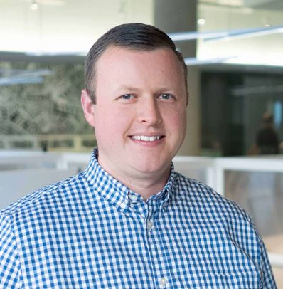 A professional headshot of a smiling man in a blue checkered shirt standing in an office environment for the team page.