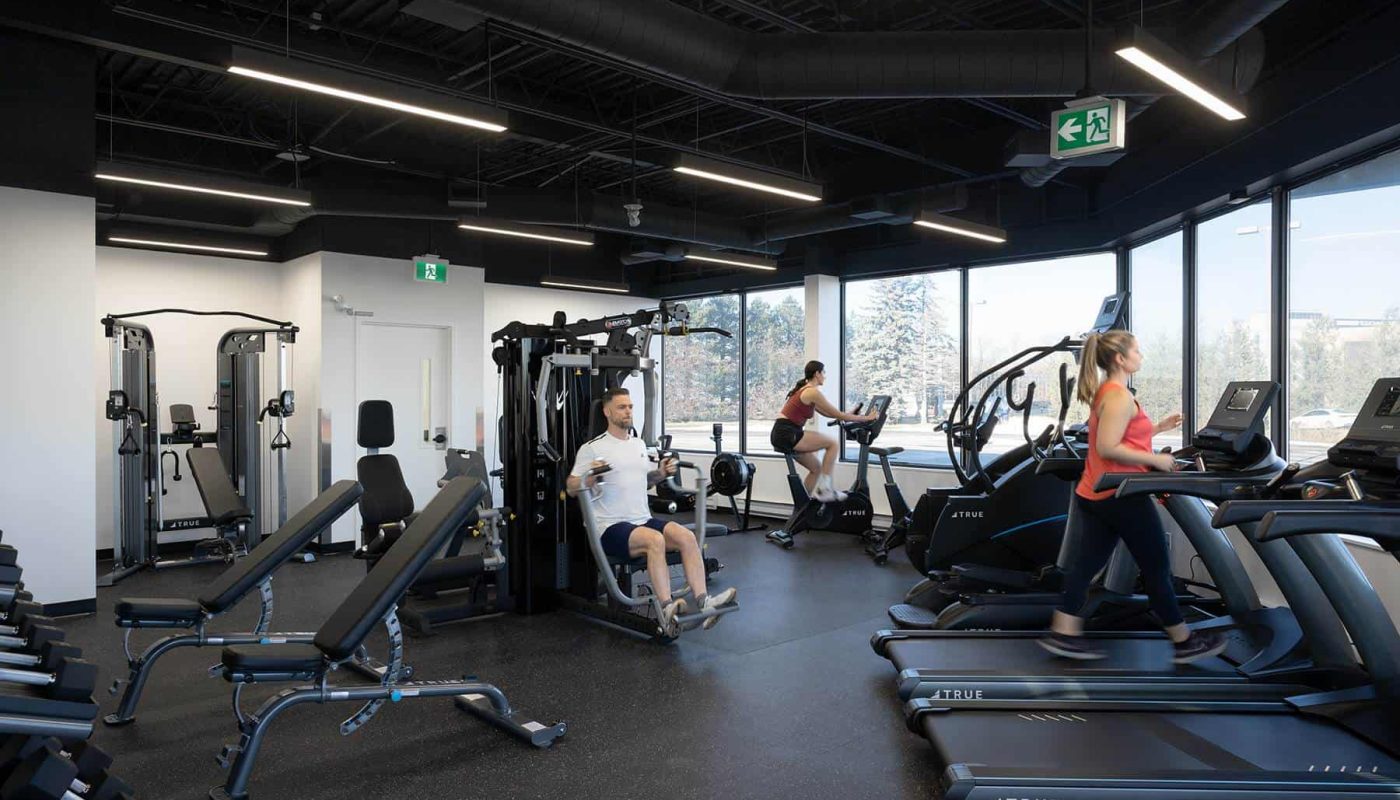 Interior of a modern gym with various exercise machines and people working out, including treadmills and weight stations, redesigned for strategic workplace design to enhance workplace wellbeing.