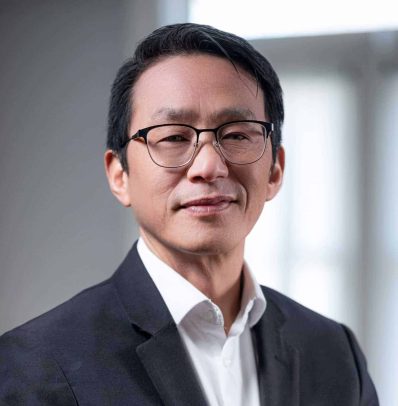 Portrait of a middle-aged Asian man wearing glasses, a black blazer, and a white shirt, smiling slightly in an office setting on the new Teams page.