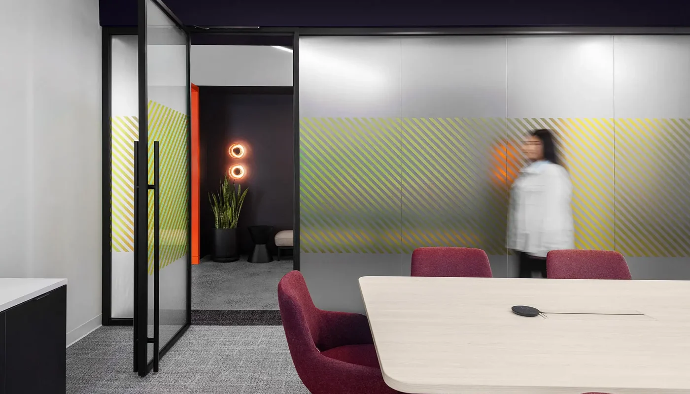 A person in motion walks by a glass partition in a modern office space. The office features a meeting room with a light wood table, red chairs, and decorative lighting.