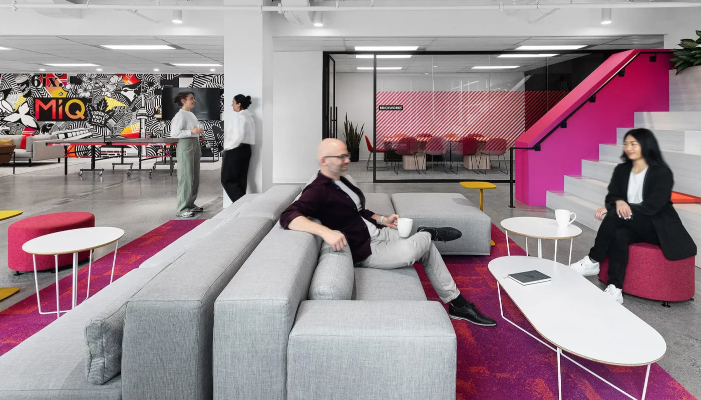 Modern office lounge with grey seating, pink accents, and three people sitting and talking. Two people standing in the background near a brightly decorated wall with "MiQ" text.