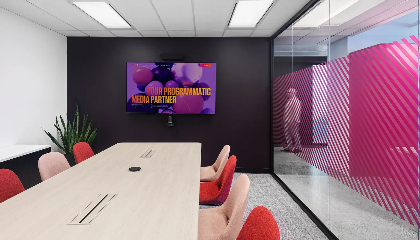 A modern conference room with a long wooden table, red chairs, a large screen displaying "Your Programmatic Media Partner," and glass walls with diagonal pink stripes.