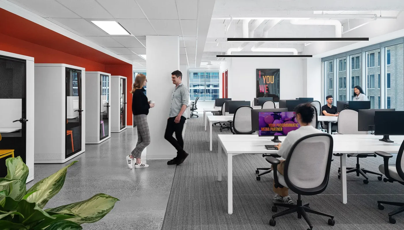 A modern office space with people working at desks and interacting. There are partitioned workstations, large windows, and overhead lighting. A plant is in the corner. One monitor screen is visible.