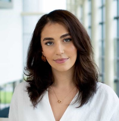 A professional portrait of a woman with shoulder-length brown hair, wearing a white blouse, featured on the new teams page in an office setting.