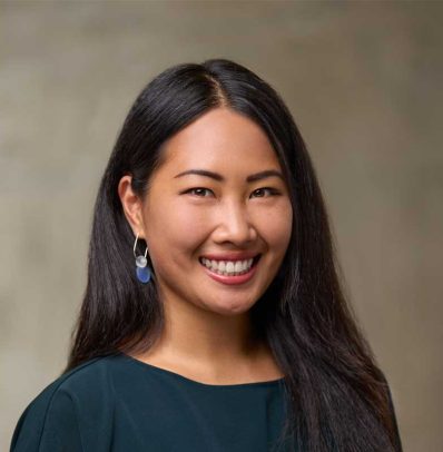 Portrait of a smiling Asian woman with long black hair, wearing a dark green blouse and blue earrings, against a neutral background for the Teams Page.